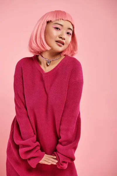 Игривая азиатская женщина с жемчужным ожерельем в ярком свитере, милая поза на розовом фоне — стоковое фото