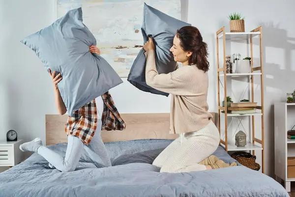Una mujer, madre, sostiene dos almohadas mientras está sentada en una cama, disfrutando de un momento de diversión en casa con su hija. - foto de stock