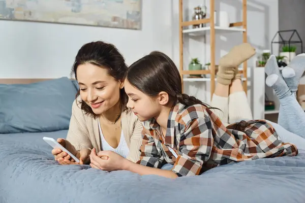 Una madre y una hija comparten un momento tierno mientras miran un teléfono celular mientras están acostadas en una cama en casa. - foto de stock