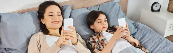 Женщина и девушка лежат на кровати, сосредоточенные на своих мобильных телефонах, наслаждаясь временем вместе дома. — стоковое фото