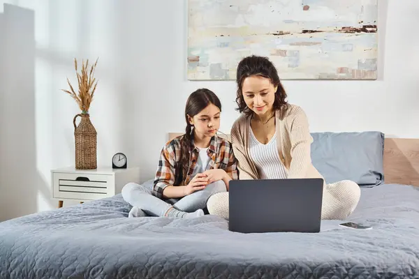 Dos mujeres sentadas en una cama, absortas en una pantalla de computadora portátil, compartiendo un momento de tiempo de calidad en casa. - foto de stock