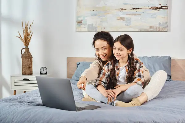Mother and daughter sit on a bed, engrossed in a laptop screen, engaging in quality time together at home. — Stock Photo