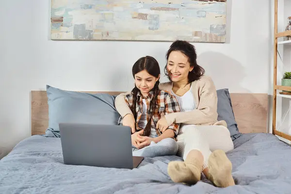 Una madre y una hija se sientan de cerca en una cama, enfocadas en una pantalla de computadora portátil con interés y conexión. - foto de stock