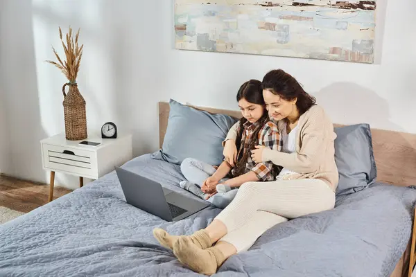 Madre e hija sentadas en una cama, absortas en la pantalla de un portátil, compartiendo un momento especial juntas. - foto de stock