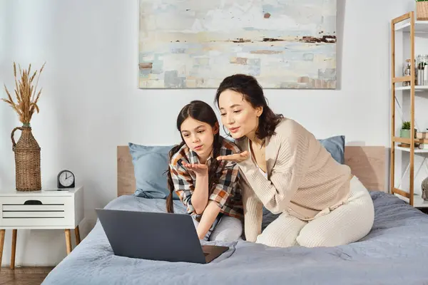 Una mujer y una chica comparten un momento tierno en una cama mientras miran juntos a la pantalla de un ordenador portátil. - foto de stock