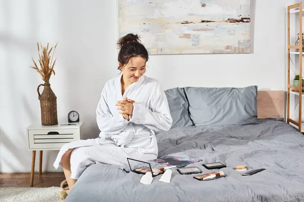 Une femme brune en robe blanche s'assoit sur un lit, entourée de cosmétiques, dans sa routine matinale. — Photo de stock
