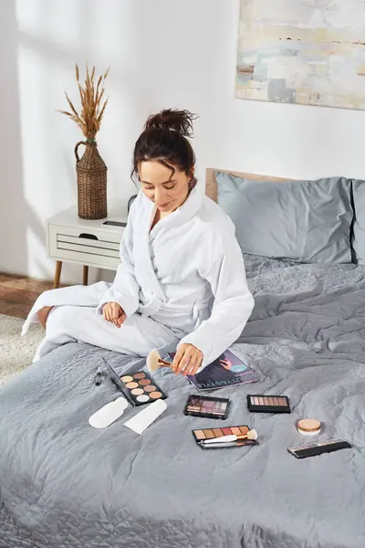 Une femme brune en peignoir blanc s'assoit sur un lit dans une chambre, appliquant du maquillage et entourée de cosmétiques le matin. — Photo de stock