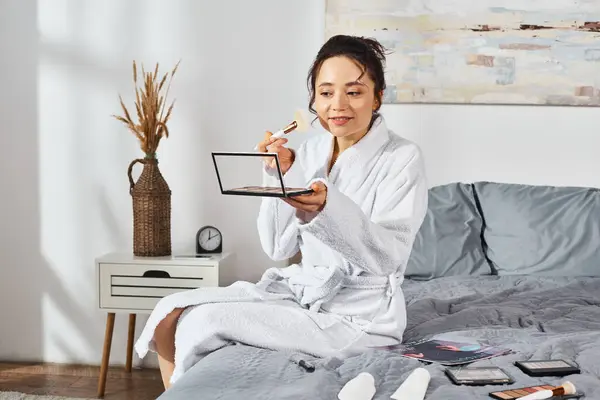 Una donna bruna in accappatoio bianco si siede su un letto con un ombretto, circondata da cosmetici, applicando il trucco al mattino. — Foto stock