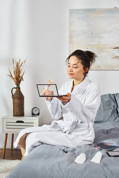 A brunette woman in a white bathrobe sitting on a bed, holding eye shadows palette and applying makeup in the morning. — Stock Photo