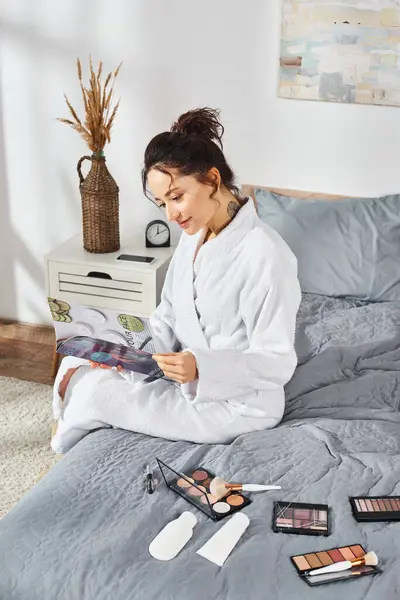 A brunette woman in a white bathrobe reading magazine on a bed in a bedroom. — Stock Photo