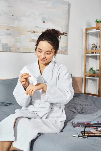 Una mujer morena sentada en una cama en un albornoz blanco, aplicando crema por la mañana. - foto de stock