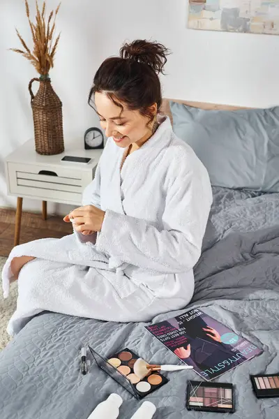 Une femme brune en peignoir blanc s'assoit sur un lit, concentrée sur sa crème tout en étant entourée de cosmétiques. — Photo de stock