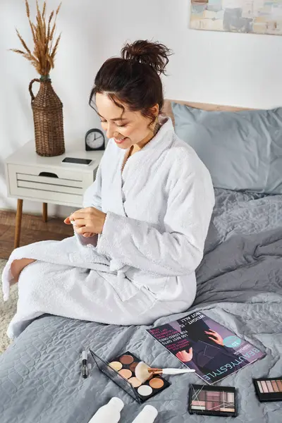 A brunette woman in a white bathrobe sits on a bed, focused on her cream while surrounded by cosmetics. — Stock Photo