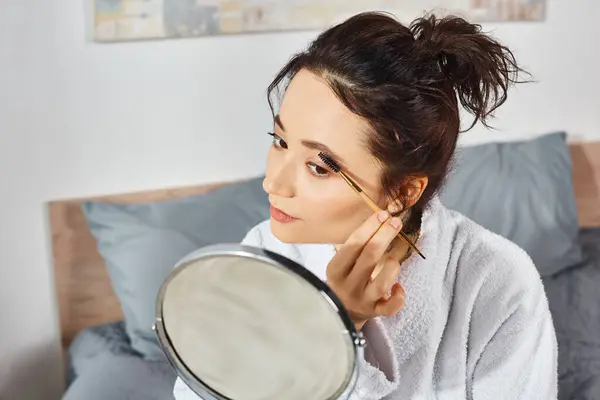Une femme brune en robe blanche s'assoit sur un lit, tenant un miroir devant son visage et appliquant du maquillage le matin. — Photo de stock
