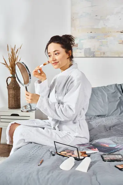 Eine brünette Frau in weißem Gewand sitzt auf einem Bett, hält sich einen Spiegel vor das Gesicht und schminkt sich. — Stockfoto