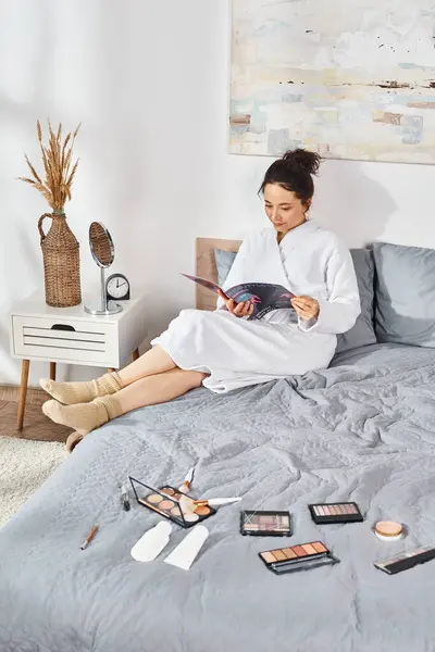 Une femme brune en peignoir blanc s'assoit sur un lit, absorbée dans un magazine, entourée de cosmétiques. — Photo de stock
