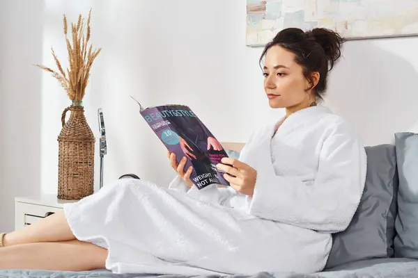 Una morena vestida con un albornoz blanco se sienta en una cama, inmersa en la lectura de una revista, rodeada de cosméticos - foto de stock