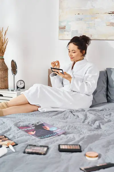 Une femme brune en peignoir blanc s'assoit sur un lit, entourée de cosmétiques alors qu'elle applique du maquillage. — Photo de stock