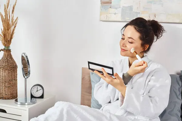 A brunette woman in a white bathrobe sits on a bed, apply makeup in the morning. — Stock Photo