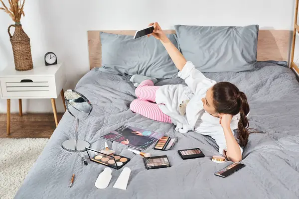 Une préadolescente brune en peignoir blanc entourée de cosmétiques et de maquillage tout en étant allongée sur un lit encombré. — Photo de stock