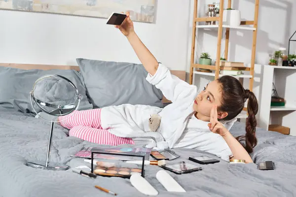 Une préadolescente brune en peignoir blanc allongée sur un lit avec un téléphone portable dans les mains, perdue dans la pensée. — Photo de stock