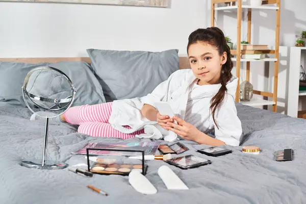 A preteen brunette girl in a white bathrobe is surrounded by cosmetics while laying on a bed. — Stock Photo