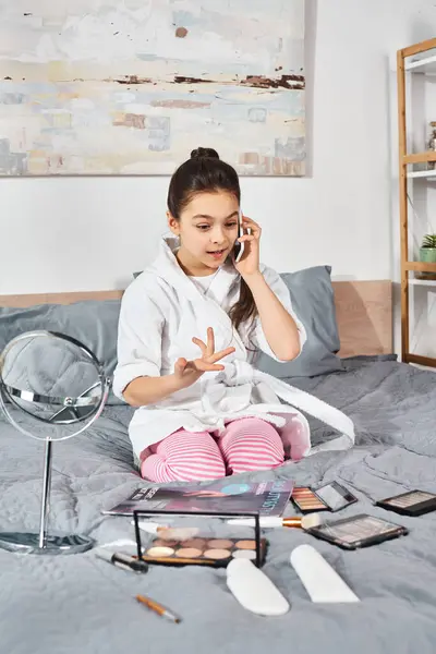Una niña preadolescente en un albornoz blanco se sienta en una cama charlando en su teléfono celular. - foto de stock