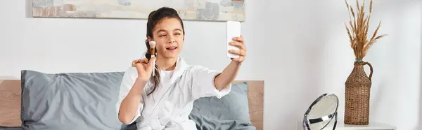 A brunette preteen girl in a white bathrobe sits on a bed holding a cell phone. — Stock Photo