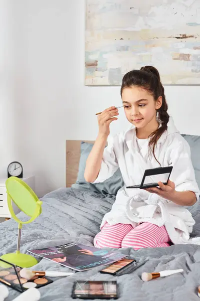 A brunette preteen girl in a white bathrobe sits on a bed, applying makeup to her face — Stock Photo