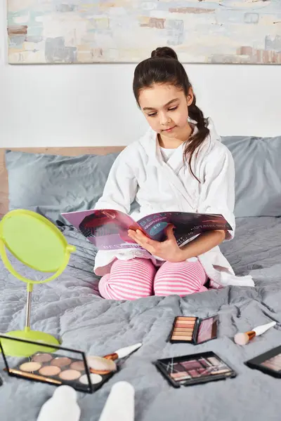 A brunette preteen girl in a white bathrobe sits on a bed, immersed in a magazine, surrounded by cosmetics. — Stock Photo