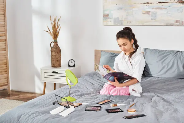 A serene scene of a brunette preteen girl in a white bath robe sitting on a bed, engrossed in a magazine with cosmetics nearby. — Stock Photo