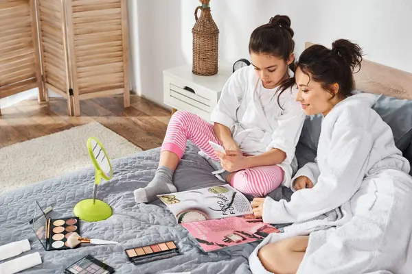 A brunette mother and daughter dressed in white bath robes sitting and bonding on top of a cozy bed. — Stock Photo