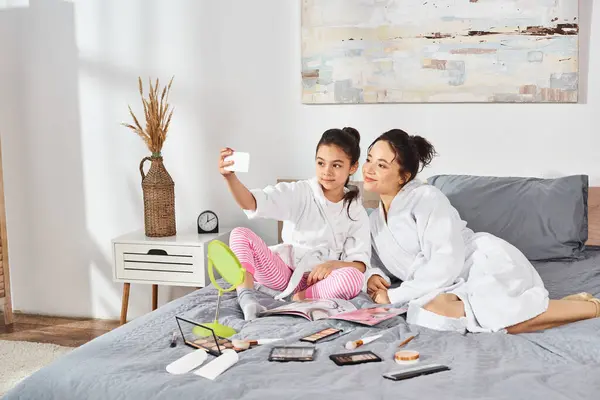 Madre e hija en batas blancas se sientan serenamente juntas en la cama, compartiendo un momento pacífico de unión. - foto de stock