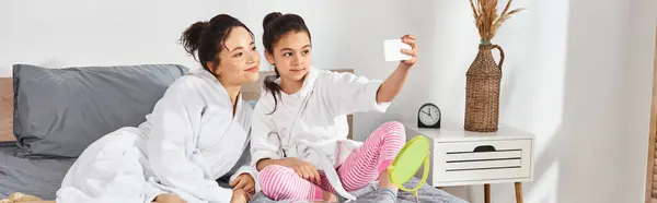Une mère brune et sa fille en peignoirs blancs assis gracieusement sur un lit dans un moment serein, prenant selfie — Photo de stock