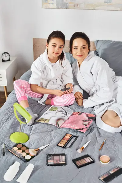 A brunette mother and daughter, both in white bath robes, sitting together on top of a cozy bed. — Stock Photo