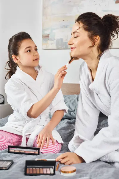 A brunette mother and her daughter, both wearing white bathrobes, sit together on a cozy bed, sharing a tender moment. — Stock Photo
