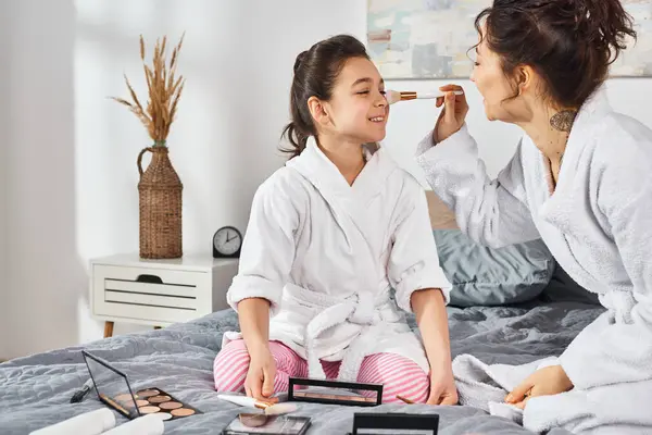 A brunette mother and daughter in white bath robes sit comfortably on a bed, applying makeup — Stock Photo