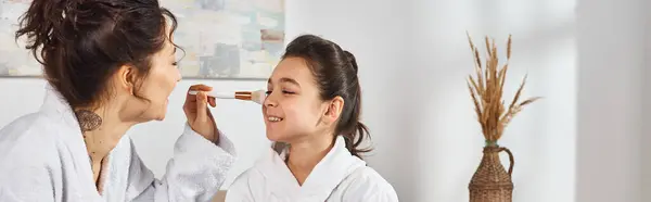 A brunette mother in a white bathrobe is delicately applying face powder on her daughters face while sitting on a bed. — Stock Photo