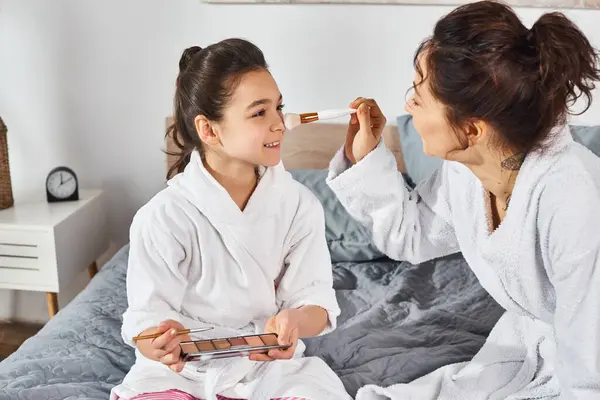 Una madre morena se sienta junto a su hija en una cama, ambas vistiendo batas blancas, compartiendo un momento especial juntos. - foto de stock