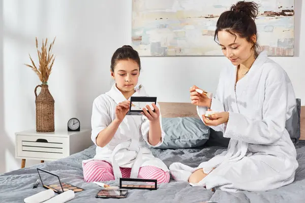 Une mère brune et sa fille en peignoirs blancs assises sur un lit, concentrées sur une palette avec maquillage — Photo de stock
