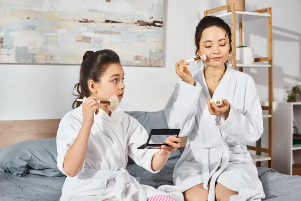 A brunette mother and daughter sit peacefully on a bed, wrapped in white bath robes, applying makeup — Stock Photo