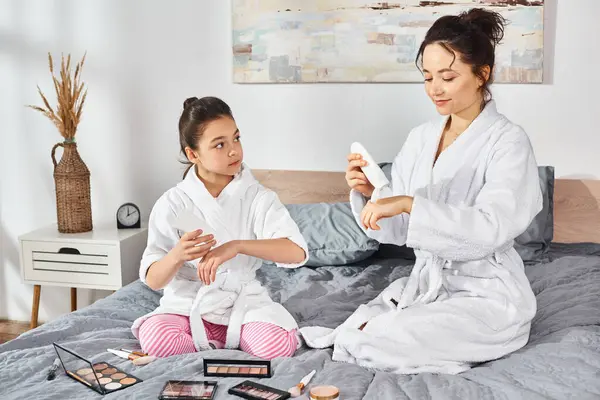 A brunette mother and her daughter wearing white bath robes are sitting together on a bed, applying cream on hands — Stock Photo