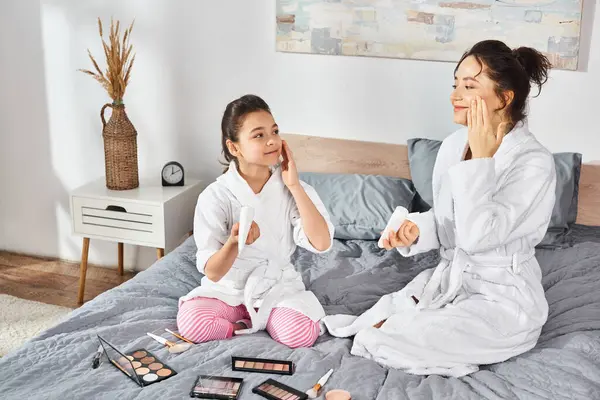 A brunette mother and daughter in white bath robes sit peacefully on a bed, applying cream, sharing a tender moment together. — Stock Photo