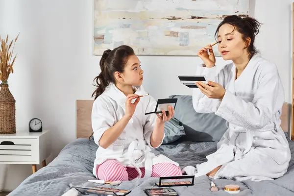 A brunette mother and daughter in white bath robes sit on a bed applying makeup together. — Stock Photo