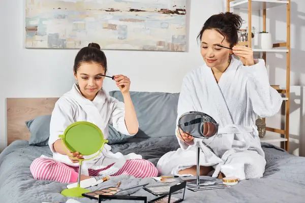 A brunette woman brushes her hair while her daughter sits on a bed, both dressed in white bath robes, applying makeup — Stock Photo