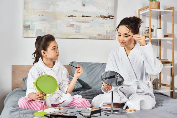 A brunette mother and daughter in white bath robes sit together on a cozy bed, sharing a tender moment, applying makeup — Stock Photo