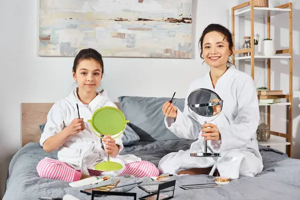 A peaceful scene of a brunette mother and daughter in white bath robes sitting comfortably on top of a bed, applying makeup — Stock Photo
