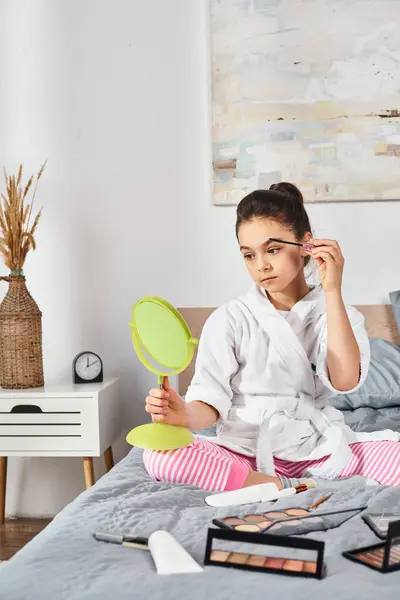 A girl with brunette hair sitting on a bed, holding a green mirror and applying mascara. — Stock Photo