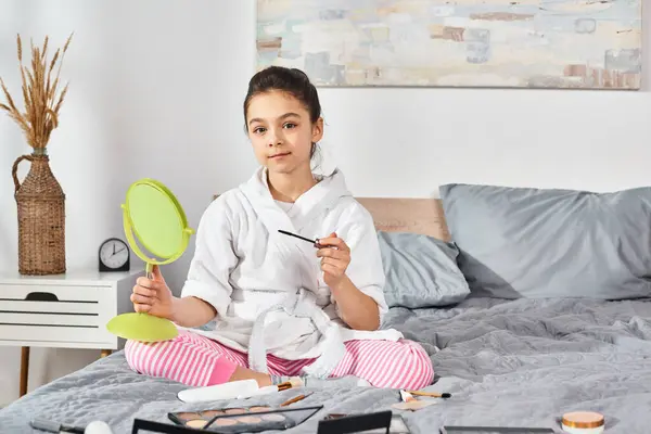 A girl in a white bath robe sits on a bed, holding a mirror, expressing curiosity and innocence. — Stock Photo