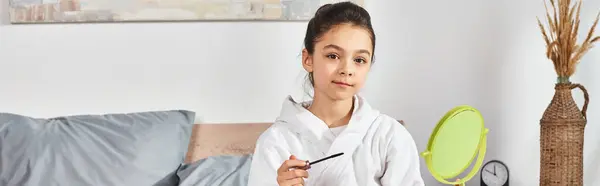 A girl with brunette hair, in a white bath robe, sitting on a bed, holding a mascara. — Stock Photo